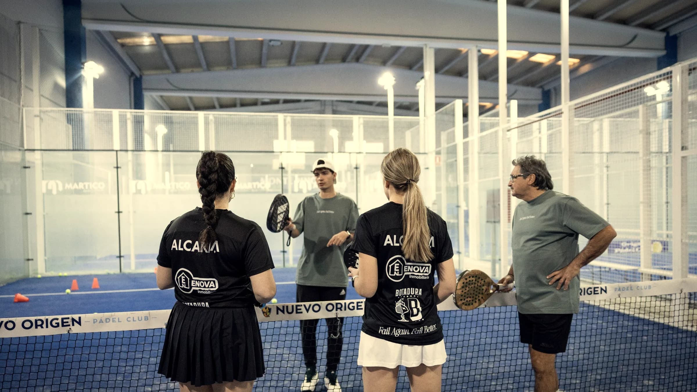A coach explains a drill to two players, seen from behind, at the net of an indoor court.