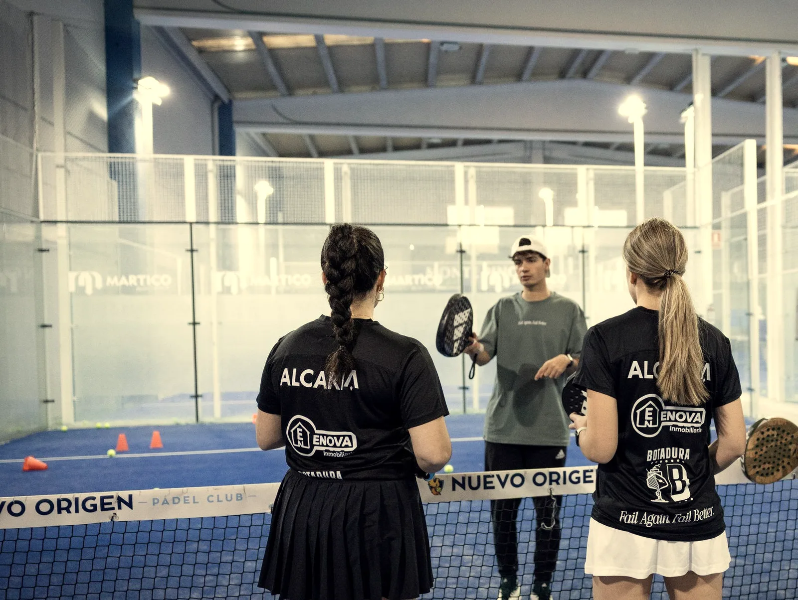 A coach explains a drill to two students, seen from behind, at the net of an indoor court