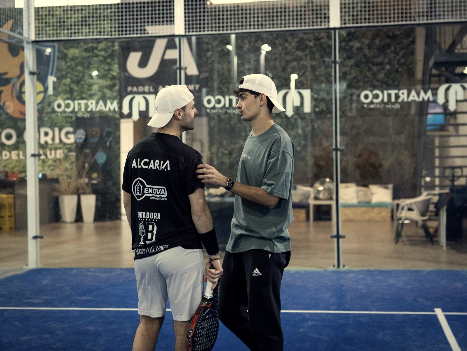 A coach in a white cap and green T-shirt talks closely to a student seen from behind, hand on his shoulder, on an indoor court
