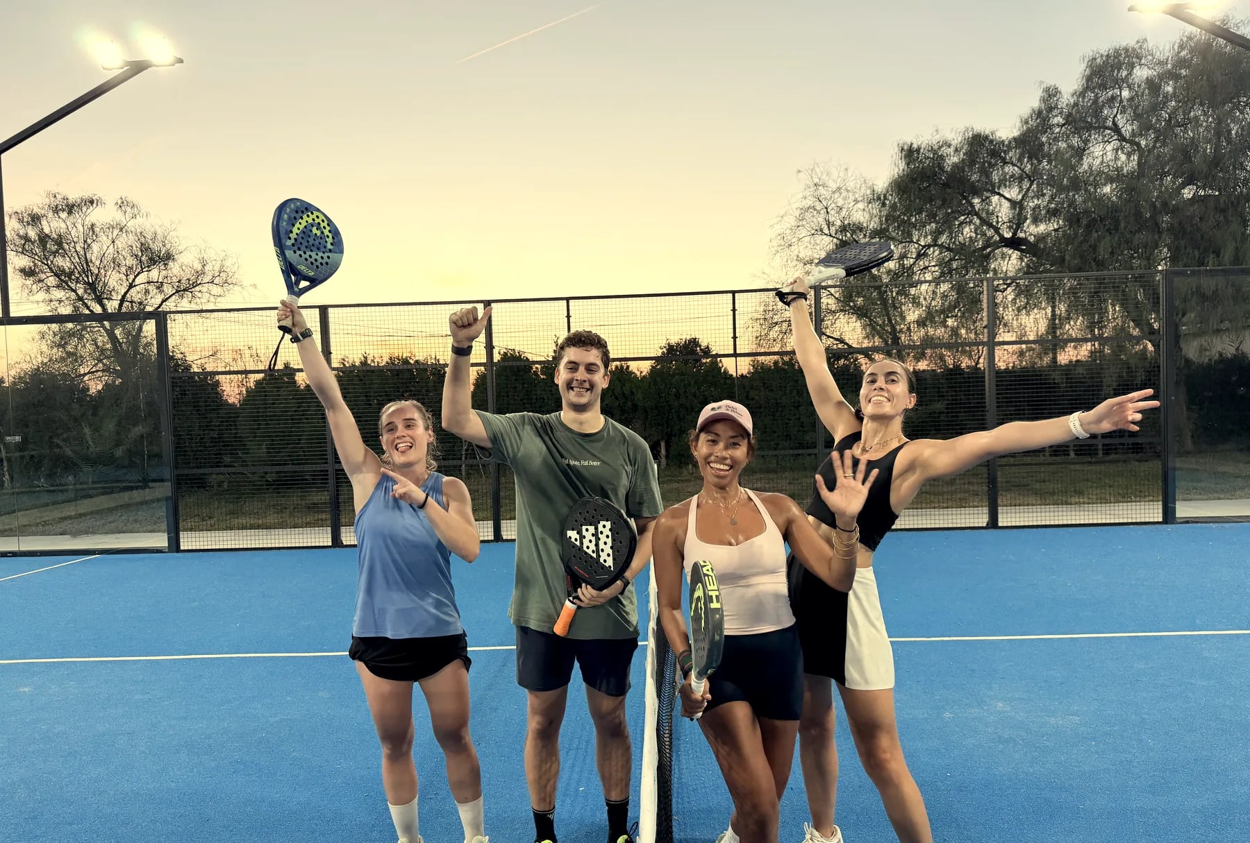Four players celebrate together at the net of a court at sunset, arms raised