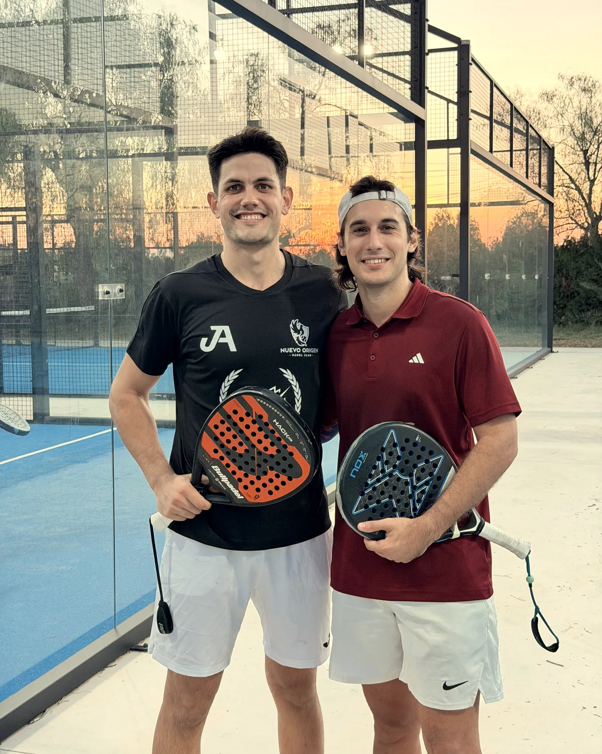 Two players smile holding their rackets at sunset: one in the academy shirt, the other in a maroon polo