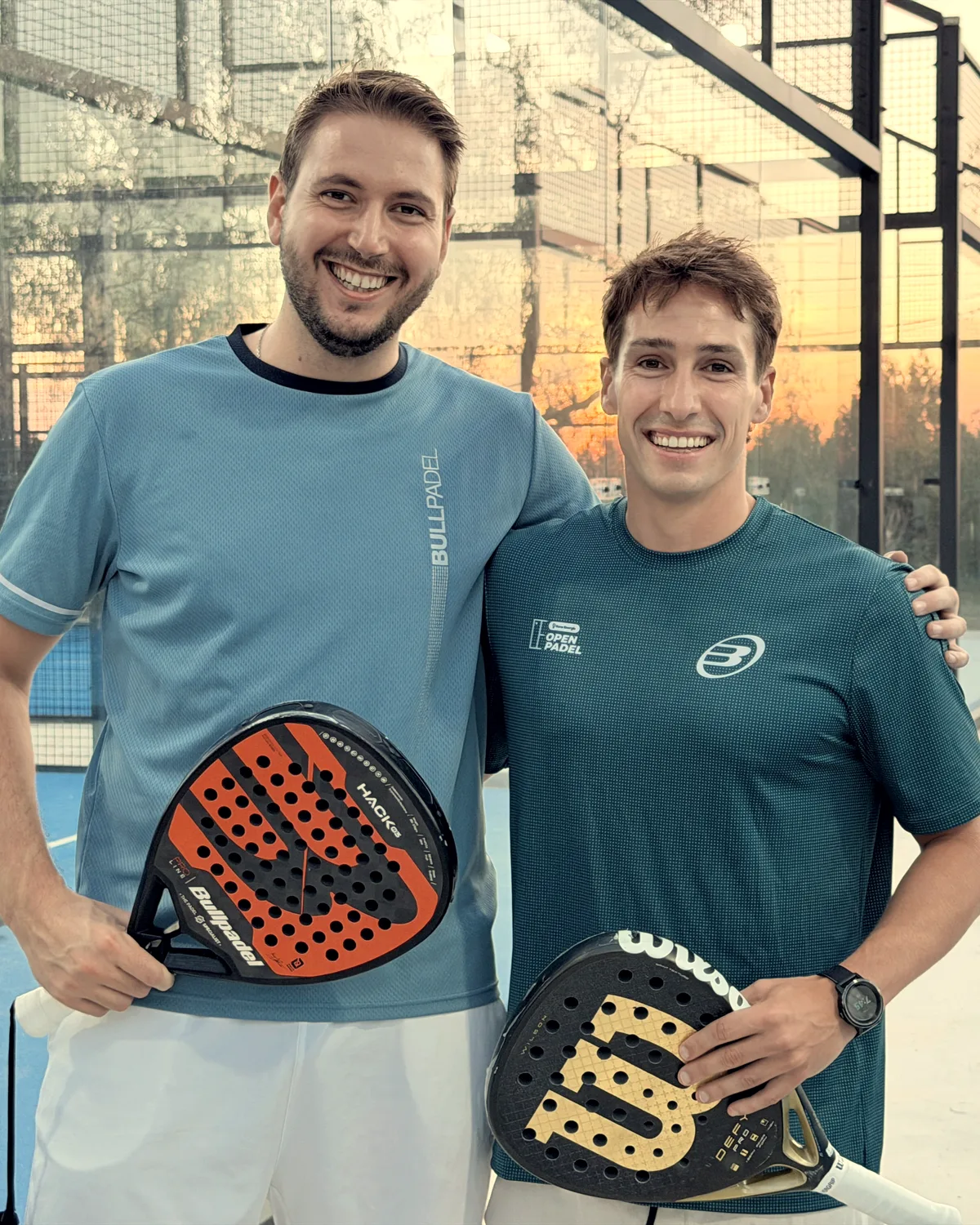 Two players smile arm in arm in the walkway between courts, the sunset behind them