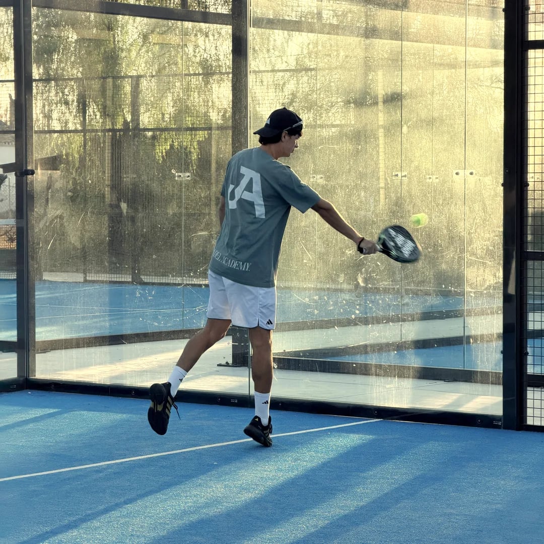 A player, seen from behind, hits the ball by the glass of an outdoor blue court