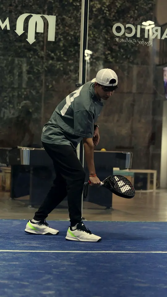 A JA coach in a white cap lowers his racket to a low ball on a blue indoor court.