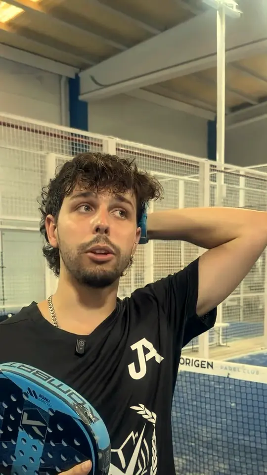 A JA coach talks with one hand behind his head and his racket in the other, beside an indoor court.