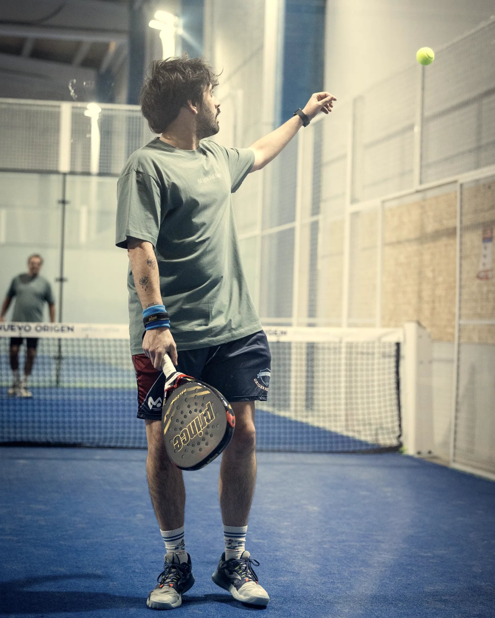 A curly-haired player in a green T-shirt tosses the ball with his left hand while holding the racket in his right, on an indoor court