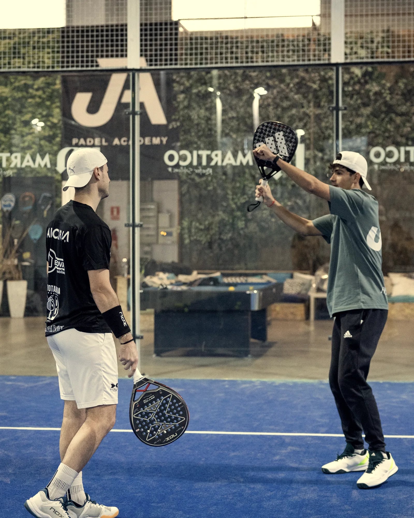 A coach in a white cap shows the stroke with raised arms to a player watching him, on an indoor court