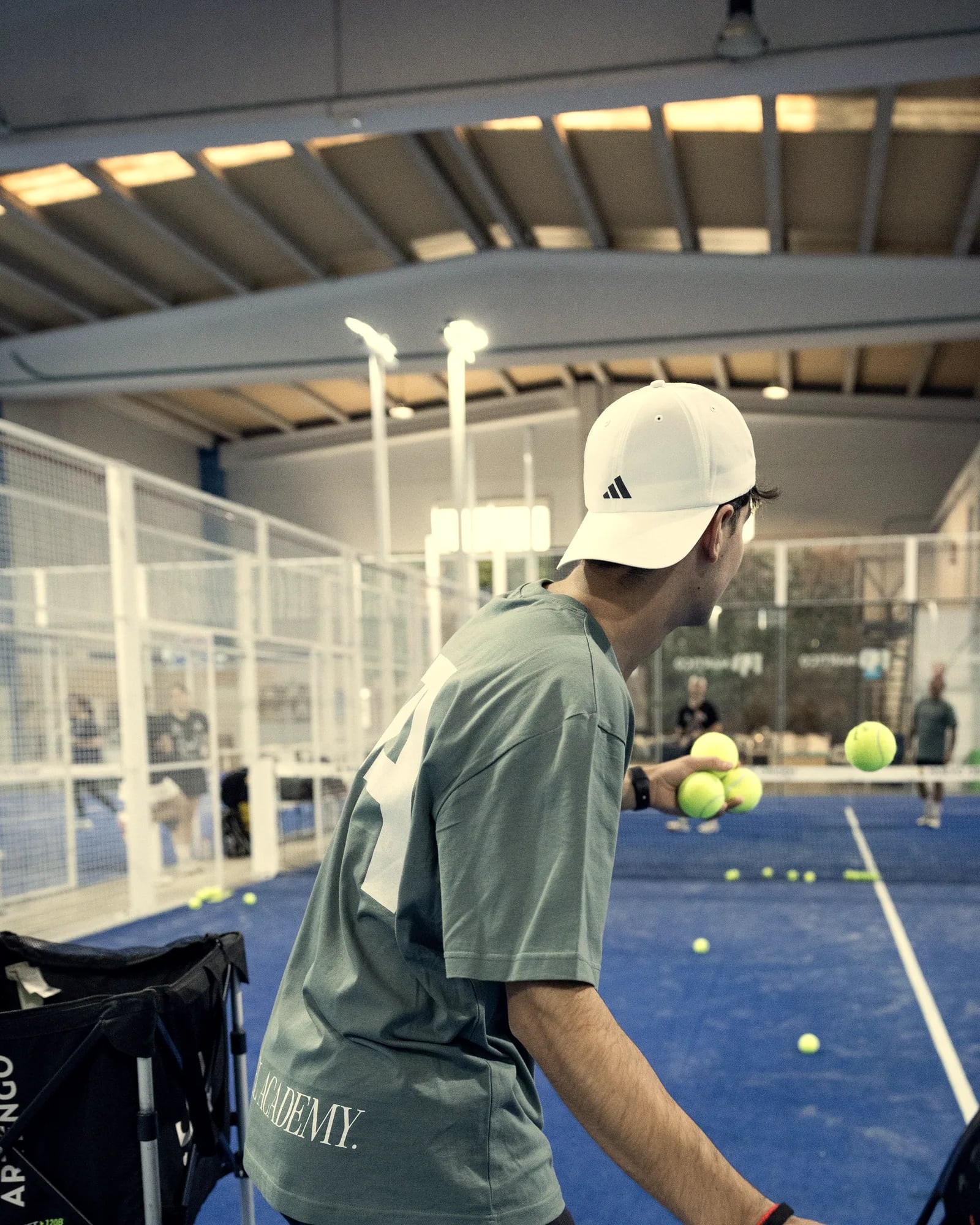 A coach feeds balls from the basket on a blue court covered in balls, under the wooden roof of the Origen Padel Club hall